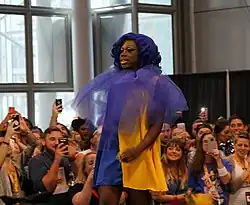 Porträtfoto einer stehenden Bob the Drag Queen. Sie trägt ein dunkelblau-gelbes Kleid und ein lilafarbenes, durchsichtiges Stofftuch um den Hals. Daneben trägt sie gelben Lippenstift, aufgemalte Augenbrauen, blau-grün-silbernen Lidschatten, Wimperntusche mit kleinen Diamanten an den unteren Wimpern und eine dunkellilane Perücke. Im Hintergrund sind ein paar Fenster, eine graue Wand und eine niedrige schwarze Trennwand zu sehen. Zudem stehen im Hintergrund mehrere Personen, von denen einige Smartphones hochhalten.