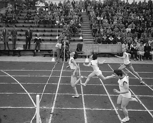 Schwarzweißfoto vom Zieleinlauf mit vier Läuferinnen, Bronnsack und Langbein auf Platz 2 und 3 in weißen Hosen und weißen Trikots, Ort und Kuyk auf 1 und 4 in vermutlich orangenen Hosen und weißen Trikots. Im Hintergrund auf Höhe der Ziellinie sitzen Fotografen.