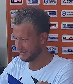 Damien Joly sitzt in einem weißen T-Shirt an einem Tisch und gibt Autogramme, wobei der Tisch in diesem Bildausschnitt nicht zu sehen ist.