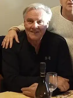 Damir Polić sitzt mit schwarzem Hemd oder Pullover an einem Tisch, vor sich eine Flasche Wein und neben sich eine Frau, die ihm den Arm auf die Schulter legt.