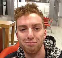 Von Daniel Wallace ist nur der Kopf zu sehen und eine schwarzweiße Kapuze im Nacken. Er befindet sich in einem Vortragssaal, im Hintergrund sind orangefarbene Stühle und eine weiße Flipchart zu erkennen.