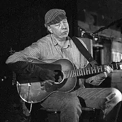 Dave "Snaker" Ray 1993 beim Auftritt im "The Cabooze", Minneapolis, mit Gitarre sitzend, in längs gestreiftem Hemd und mit Kappe