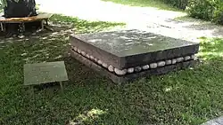 Das Bild zeigt eine metallische Gedenktafel neben zwei aufeinander gelegten Steinplatten. Zwischen den Steinplatten befinden sich kleinere Steine.