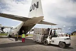 Vor einem Flugzeugheck entlädt ein Gabelstaplerfahrer Paletten mit Paketen. Die Fahrzeuge sind mit UN beschriftet.