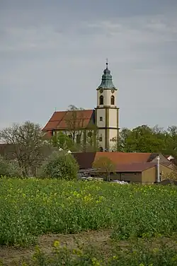 Die relativ kleine weiße Kirche mit rotem Ziegeldach aus der Ferne, im Vordergrund Rapsfelder.
