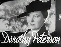 Im Auto sitzenden Frau im mittleren Alter im Seitenprofil in die Kamera schauend in heller Bluse und mit schwarzen Hut in schwarzweiß mit ihrem horizontalen Namenszug im unteren Bildbereich