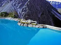 Blick aus Richtung einer Talsperre auf eine mehrgeschossige Berghütte, gebaut auf einem Felsvorsprung in Verlängerung der rechts daneben befindlichen Staumauer der Talsperre; im Hintergrund links ein Bergmassiv aus puren Fels; rechts der Talabgrund jenseits der Staumauer