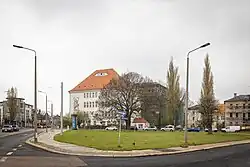 Foto eines vierstöckigen, breiten Gebäudes mit hohem Dach hinter einer Grünfläche. Auf der linken Seite des Gebäudes ist ein Fassadenbild zu sehen, das Rübezahl zeigt,