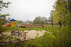 Fotografie eines Geländes mit Sandgrube, in der ein buter Plastikrutschenturm steht. Äste einer Hecke und von Bäumen mit jungem Grün ragen von beiden Seiten und von unten in das Bild