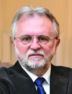 Bebrillter älterer Mann von vorn ab Schulterblatt im dunklen Jacket, weißem Hemd und dunkler Krawatte in die Kamera schauend in Farbe.