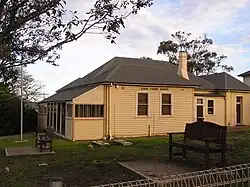 Das Foto eines weißlich-gelben Holzhauses mit einem grauen Dach. Das Haus ist sehr verwinkelt gebaut, besitzt einen Schornstein und ist mit EDEN COURT HOUSE beschriftet. Vor dem Haus stehen auf einer Wiese zwei Holzbänke sowie ein Fahnenmast, an dem allerdings keine Fahne weht.