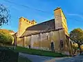 Westseite einer perigordinischen Wehrkirche im Abendlicht mit blauem Himmel und Teilen des Dorfkerns als Hintergrund