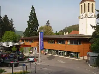 Gemeindezentrum von Westen, mit den Türmen der beiden römisch-katholischen Kirchen im Hintergrund. Im Erdgeschoß links die Kaffee-Konditorei rechts die Bibliothek.