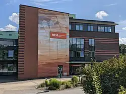 Ein Backstein- und Glasgebäude des Herzogin Elisabeth Hospitals. Links bedeckt ein großes, abstraktes Wandgemälde in Braun-, Orange- und Weißtönen eine Backsteinfassade, auf der sich das orangefarbene Logo „HEH Herzogin Elisabeth Hospital“ befindet. Rechts ist der Eingang zum „Ärztehaus“.