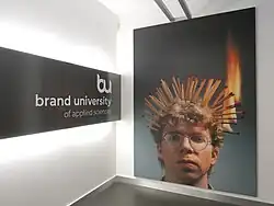 Eingangsbereich der Brand University mit Fotografie einer kreativen Arbeit: ein junger Mann mit Streichhölzern in den Haaren. Einige von ihnen brennen.