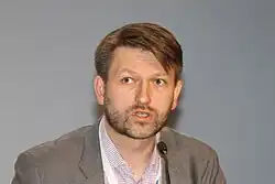 Foto eines braunhaarigen Mannes mit Vollbart, der in ein Mikrofon spricht