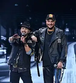 Foto zweier in Schwarz gekleideter Männer, der linke singt in ein Mikrofon