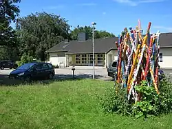 Auf der Wiese steht ein etwa drei Meter hohes, ungeordnetes Bündel armdicker Holzpfähle, die mit farbenfrohen Mustern bemalt sind. Das Arrangement erinnert an ein Bündel Mikadostäbe vor dem Fallenlassen.