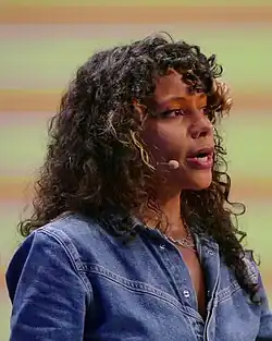 Seitliches Porträt einer BIPoC Frau mit Locken, die leicht nach oben blickt. Sie steht vor einem gelb-orangenen Hintergrund und trägt ein Oberteil aus Jeansstoff.