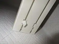 Ende einer Vorhangschiene mit Aussparungen zum Ein- und Ausfädeln der Gardine und einem Gardinenfeststeller (links)