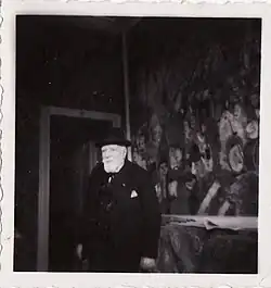 Schwarz-Weiß-Fotografie des alten James Ensor in Anzug, Hut und mit weißem Bart in seinem Haus. An der Wand ist ein Ausschnitt des Gemäldes „Der Einzug Christi in Brüssel“ zu sehen.