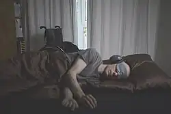 Eine an ME/CFS erkrankte Person liegt auf der Seite in einem Bett und ist mit einer braunen Decke zugedeckt. Die Person trägt ein graues T-Shirt, eine Schlafmaske und einen Gehörschutz. Der Raum ist mit Vorhängen abgedunkelt. Neben dem Bett steht ein schwarzer Rollstuhl.