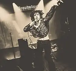 Das in schwarz-weiß gehaltene Foto zeigt den Musiker Ethan Bortnick auf seiner Tournee im Nachtclub „The Troubadour“ in West Hollywood. Der Musiker hat schwarze Locken, trögt ein dunkler Netzhemd sowie eine hüfthohe Jeans. In der rechten Hand hält er ein Mikrofon in der Hand, die linke streckt er in die Luft. Hinter Bortnick stehen ein Mikrofonständer, ein Klavier mit Hocker, ein Verstärker sowie ein weiterer Musiker.