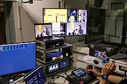 Verschiedene technische Geräte mit Monitoren auf einem grauen Tisch, am unteren rechten Rand des Bildes sind die Hände einer Veranstaltungstechnikers zu sehen, eine davon greift eine Art Joystick an einem Mischpult