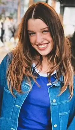 Ever Anderson (2024) hat lange, wellige Haare, die über die Schultern fallen und lächelndem Gesicht. Sie trägt eine blaue Jeansjacke über einem blauen T-Shirt. Im Hintergrund sind verschwommene Figuren zu erkennen, die möglicherweise andere Menschen darstellen, sowie eine städtische Umgebung. Jedoch sind die spezifischen Details im Hintergrund nicht klar erkennbar.