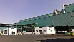 Das Bild zeigt ein grünes Industriegebäude mit zwei kleinen weißen Häusern und einigen Rohren vor sich.