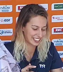 Fantine Lesaffre sitzt während einer Autogrammstunde vor einer Werbetafel. Sie trägt ein dunkelblaues Polohemd und hält in der rechten Hand einen Stift.