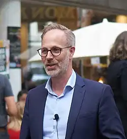 Foto eines mittelalten Mannes in einer Stadt. Er trägt graue Haare die zur Seite gekämmt sind, einen grauen Dreitagebart und eine Brille. Er scheint mit einer Person zu sprechen. Weiterhin trägt er ein dunkelblaues Sakko über einem hellblauen Hemd. Der Kragenknopf des Hemds ist geöffnet, an der Knopfleiste ist ein Mikrofon befestigt. Im Hintergrund sind unscharf verschiedene Menschen zu erkennen, die teilweise unter dem weißen Sonnenschirm eines Cafés oder Restaurants sitzen.