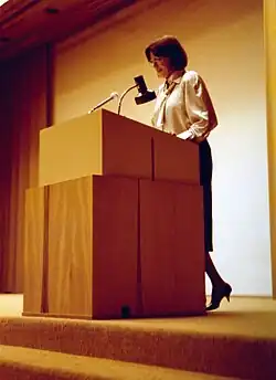 Yvonne Escher, die hinter einem hölzernen Podium steht, als ob sie eine Rede oder Präsentation hält. Das Podium hat einen befestigten Mikrofonständer sowie eine kleine Lampe, die darauf gerichtet ist. Die Person trägt eine hellfarbene Bluse kombiniert mit einem dunklen Rock und befindet sich auf einer Bühne mit Teppichboden. Der Hintergrund ist schlicht gehalten und besteht aus einer Wand mit einem Vorhang auf der linken Seite.