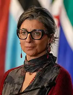 Francesca Albanese. Sie trägt ein rotes Top, einen grauen Schal, rote Ohrringe, und eine schwarze Brille mit großen Gläsern. Der Hintergrund besteht aus verschiedenen Flaggen. Die Szenerie entstand bei einer Konferenz in Bogotá 2025.