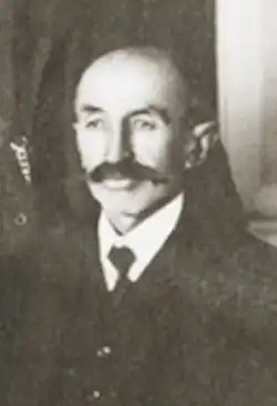 Portrait von Hoop: wenig Haare, starke, schwarze Augenbrauen, großer, schwarzer Schnäuzer, gekleidet in einen schwarzen Anzug mit weißem Hemd und Krawatte, vor unbestimmten Hintergrund