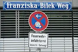 Foto eines blauen Straßenschilds mit weißer Schrift "Franziska-Bilek-Weg. Unter dem Straßenschild ist ein Parkverbotschild mit Zusatzschild "Feuerwehranfahrtszone (einschließlich Gehweg)". Hinter den Schildern sieht man geschlossene Jalousien eines Fensters.