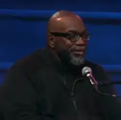 Fred Moten, ein Schwarzer Mann mit Glatze vor einem Mikrofon und dunkelblauen Vorhang im Hintergrund.