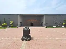 Die Gedenkstätte mit rot gepflasterten Platz und flach gehaltener Gedächtnishalle für die Zwangsarbeiter von Fuxin, im Vordergrund eine Skulptur aus einer Lore und darin zusammengedrängten Minenarbeitern