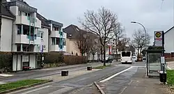 Städtische Straße (Gänsewinkel), die von mehreren Wohngebäuden gesäumt ist. Auf der linken Seite befinden sich hell gestrichene Gebäude mit Balkonen. Zwischen den Gebäuden steht eine Bushaltestelle mit einem überdachten Wartebereich und einer Sitzbank. Rechts im Bild sieht man eine weitere Bushaltestelle mit einem Schild, das die Haltestelle „Gänsewinkel“ kennzeichnet. Das Schild listet die Buslinien R30, R31 und R50 auf. Ein weißer Bus mit der Nummer 539 fährt gerade die Straße entlang.Die Straße ist von kahlen Bäumen gesäumt, was auf eine winterliche oder frühe Frühlingszeit hindeutet. Einige Autos sind ebenfalls zu sehen, sowohl geparkt als auch in Bewegung.