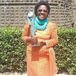 Katana Gégé Bukuru. Afrikanische Frau mit Kraushaaren in einem orangen Kleid mit einem minzgrünen Schal und einer Preis-Trophäe aus Bronze.