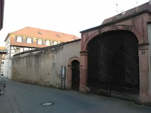 Eingangsportal und Umfassungsmauer des ehemaligen Adelshofes mit Blick entlang der heutigen Brunnengasse (im Bild links) Richtung Osten. Die als Wohnhaus restaurierte ehemalige Scheune (im Hintergrund) gehörte früher mit zum Anwesen.