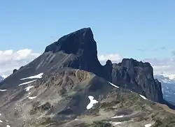 Ein zerklüfteter Berg mit seinem Hauptgipfel umgeben von einer Bergkette zur Rechten und einer linken, von Geröll bedeckten Flanke