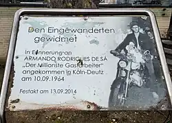 Schräg angebrachte, gedruckte Gedenktafel unter Glas oder Acrylglas, gefasst an drei Seiten in einen Rahmen aus Stahlrohr. Schlechter Zustand mit Verschmutzung und Feuchtigkeitsflecken hinter dem Glas. Aufschrift: „Den Eingewanderten gewidmet. In Erinnerung an Armando Rodrigues de Sá „Der Milionste Gastarbeiter“ angekommen in Köln-Deutz am 10.09.1964. Festakt am 13.09.2014.“ Rechts freigestelltes Foto des Arbeiters mit einem Blumenstrauß auf seinem geschenkten Motorrad.