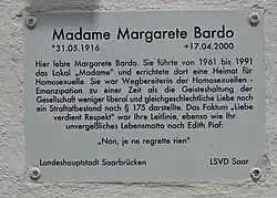 In schwarzer Schrift auf weißem Untergrund ist zu lesen: Madame Margarete Bardo *31.05.1916 +17.04.200 Hier lebte Margarete Bardo. Sie führte von 1961–1991 das Lokal „Madame“ und errichtete dort eine Heimat für Homosexuelle. Sie war Wegbereiterin der Homosexuellen. Emanzipation zu einer Zeit als die Geisteshaltung der Gesellschaft weniger liberal und gleichgeschlechtliche Liebe noch ein Strafbestand nach § 175 darstellte. Das Faktum „Liebe verdient Respekt“ war ihre Leitlinie, ebenso wie ihr unvergeßliches Lebensmotto nach Edith Piaf: Non, je ne regrette rien! Landeshauptstadt Saarbrücken LSVD Saar