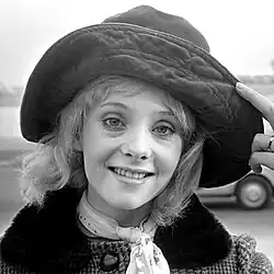 Geneviève Waïte lächelt freundlich und trägt einen großen Hut mit breiter Krempe, der aus einem weichen Material gefertigt ist. Der Hut verdeckt den oberen Kopfbereich und gibt den lockigen, hellen Haaren Raum, unter ihm hervorzuschauen. Um den Hals der Person liegt ein Schal mit einem Punktemuster. Sie trägt außerdem einen Mantel mit einem Pelzkragen und einem großen Knopf als Verschluss. Ihre rechte Hand hält den Hut an der Krempe fest, und an einem Finger ist ein Ring sichtbar.