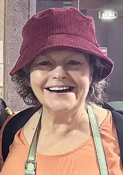 Genevieve Lemon lächelt und trägt einen roten roten Cord-Bucket-Hut. Sie hat lockiges Haar, das teilweise unter dem Hut hervorschaut, und trägt ein orangefarbenes Oberteil. Auf dem Rücken hat Genevieve Lemon einen Rucksack mit grünen Trägern. Im Hintergrund ist eine Steinwand zu sehen, und ein Schild mit der Aufschrift „EXIT“ leuchtet in weißen Buchstaben. Es gibt außerdem ein rotes Schild, das teilweise hinter ihr sichtbar ist.