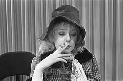 Geneviève Waïte trägt einen karierten Mantel mit einem Pelzkragen und hat welliges Haar, das bis zu den Schultern reicht. Sie hält eine Zigarette in der rechten Hand und zieht an dieser. Um den Hals trägt sie ein gemustertes Halstuch. Der Hintergrund zeigt vertikale Streifen, die möglicherweise zu einem Vorhang gehören.