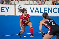 Georgina Oliva im roten spanischen Nationaltrikot führt den Ball auf blauem Kunstrasen. Rechts im Vordergrund ist der Rücken der Waliserin Xenna Hughes.