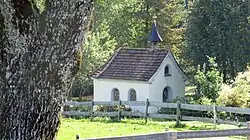 Kapelle in weiß mit Glocke auf Dach, davor ein Zaun