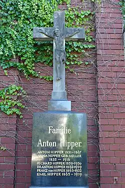 Grabkreuz mit schwarzem Sockel vor roter Ziegelwand mit Efeubewuchst. Auf dem Sockel sind in goldener Schrift sechs Namen und Sterbedaten von Mitgliedern der Familie Hipper eingelassen.